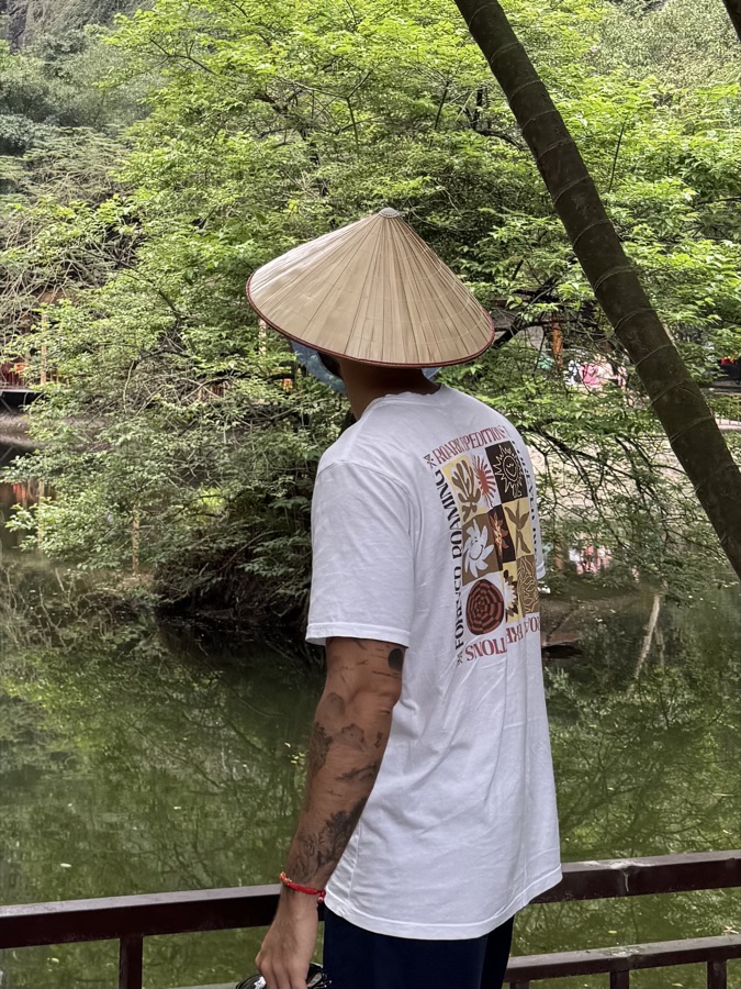 Kai Tsukimi standing by still water, face turned away beneath a woven hat